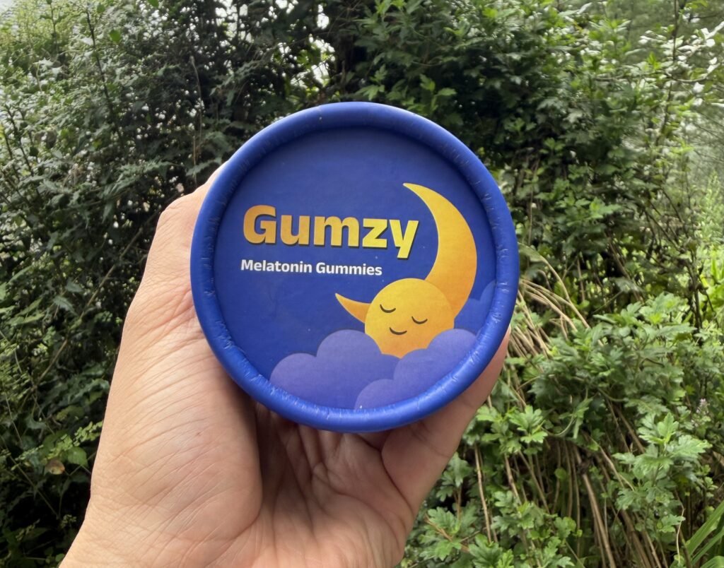 A hand holding a blue Gumzy Melatonin Gummies jar lid, featuring an illustrated sleepy crescent moon character resting on clouds.